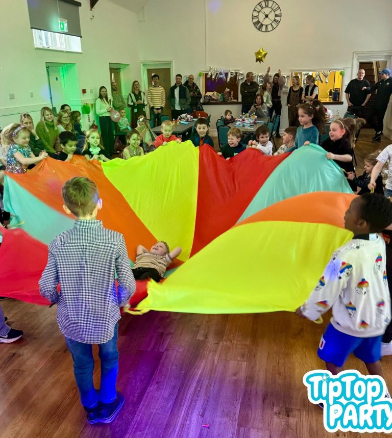 Children and adults playing with a large, brightly coloured parachute in a lively room.