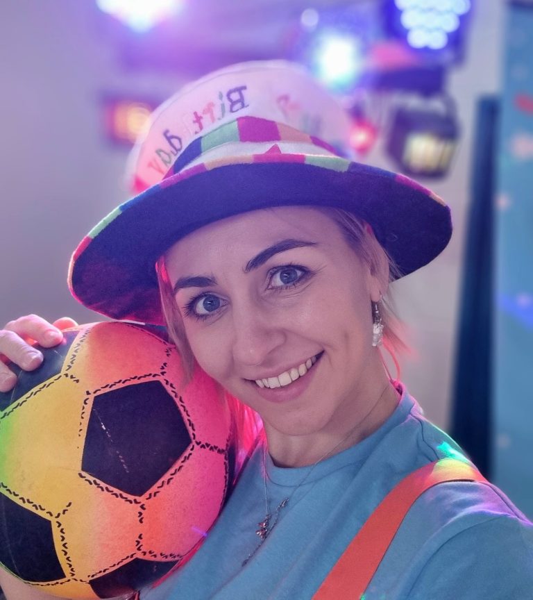 Smiling woman holding a colourful soccer ball, wearing a festive hat and bright clothing.